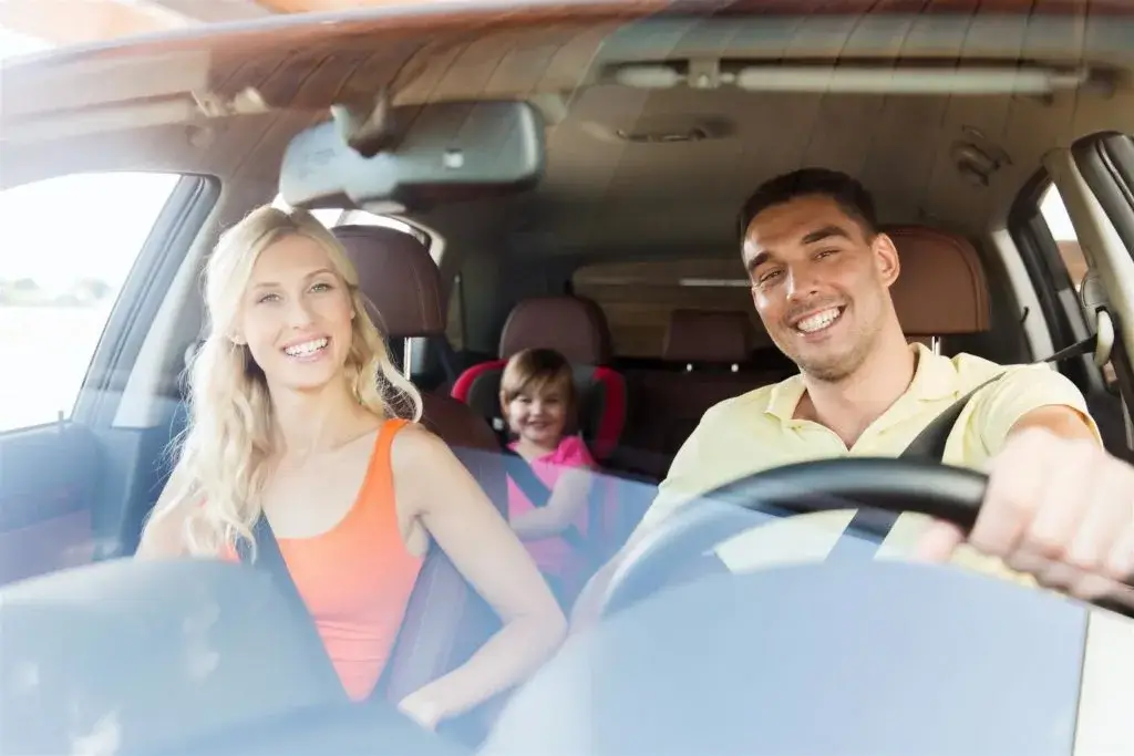 A smiling family sits in a car with their seatbelts fastened, with a child in a car seat in the back