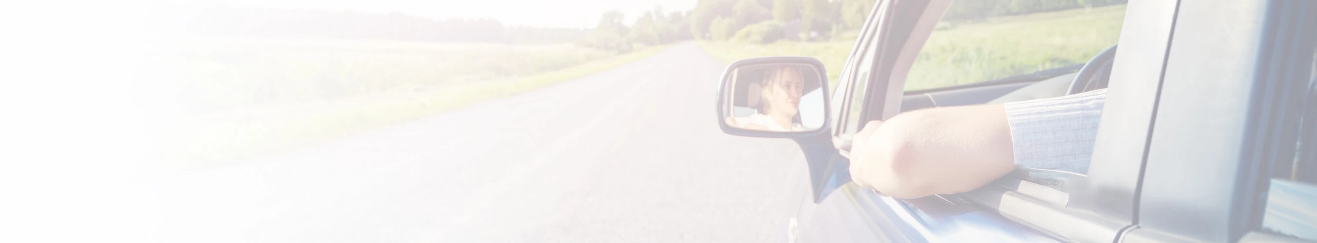 Person driving with their arm resting on the car window, looking at the road ahead with a view of the side mirror reflecting their face.