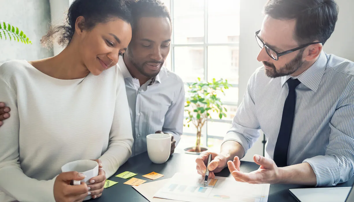 A couple consults with an insurance agent, looking at documents on the table while the agent explains details