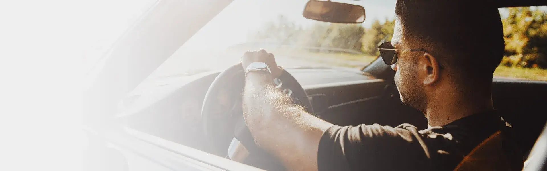 Man driving a car with one hand on the steering wheel, wearing sunglasses on a sunny day