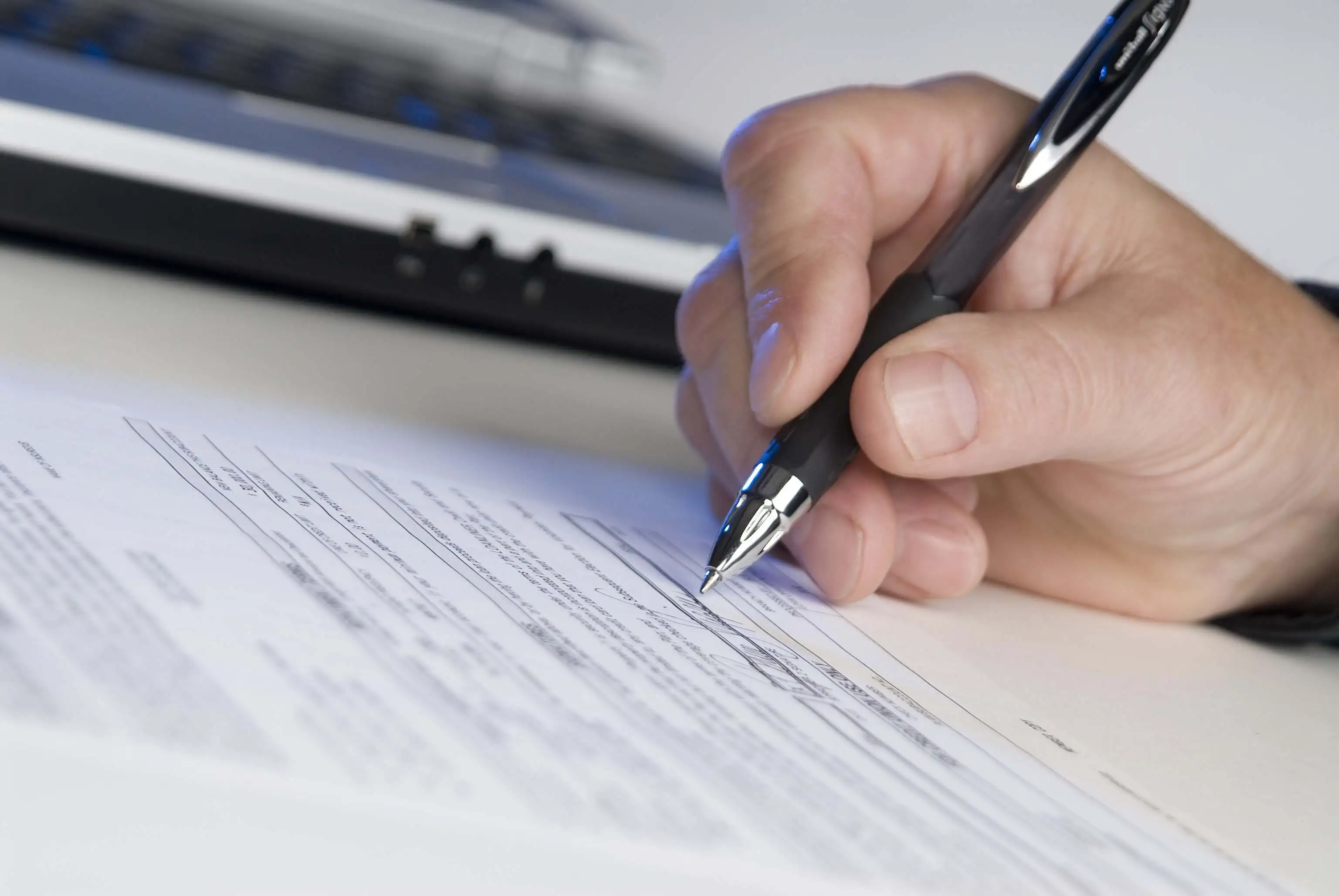 A person signing a document with a pen, with a laptop in the background.