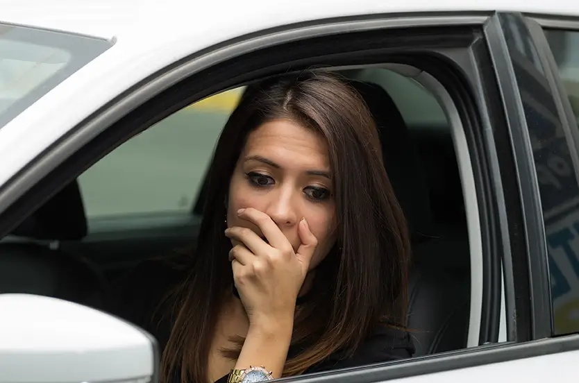 A woman in a car, looking worried, with her hand covering her mouth as she stares out the window.