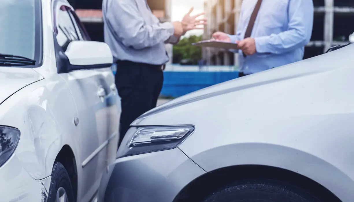 Two people in business attire examine the damage to two cars involved in a minor collision, with one person holding a clipboard.